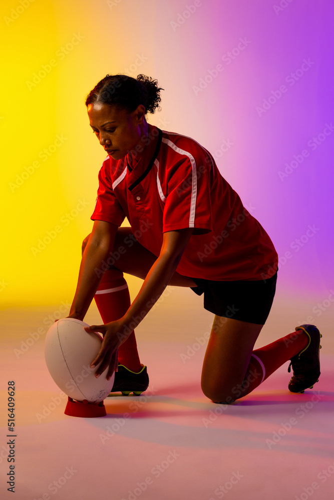 African american female rugby player crouching with rugby ball over neon pink lighting