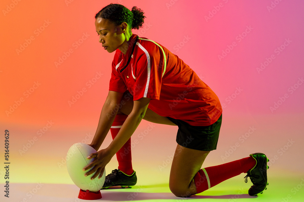 African american female rugby player crouching with rugby ball over neon pink lighting