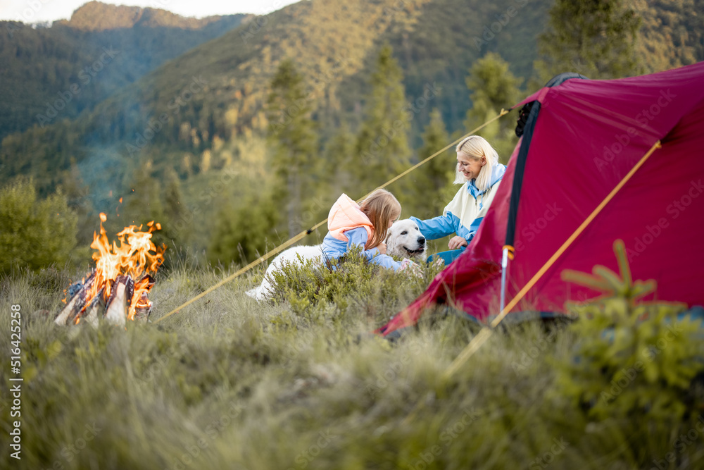 Young woman with her little girl and dog sit by the campfire, bonding together while travel on natur