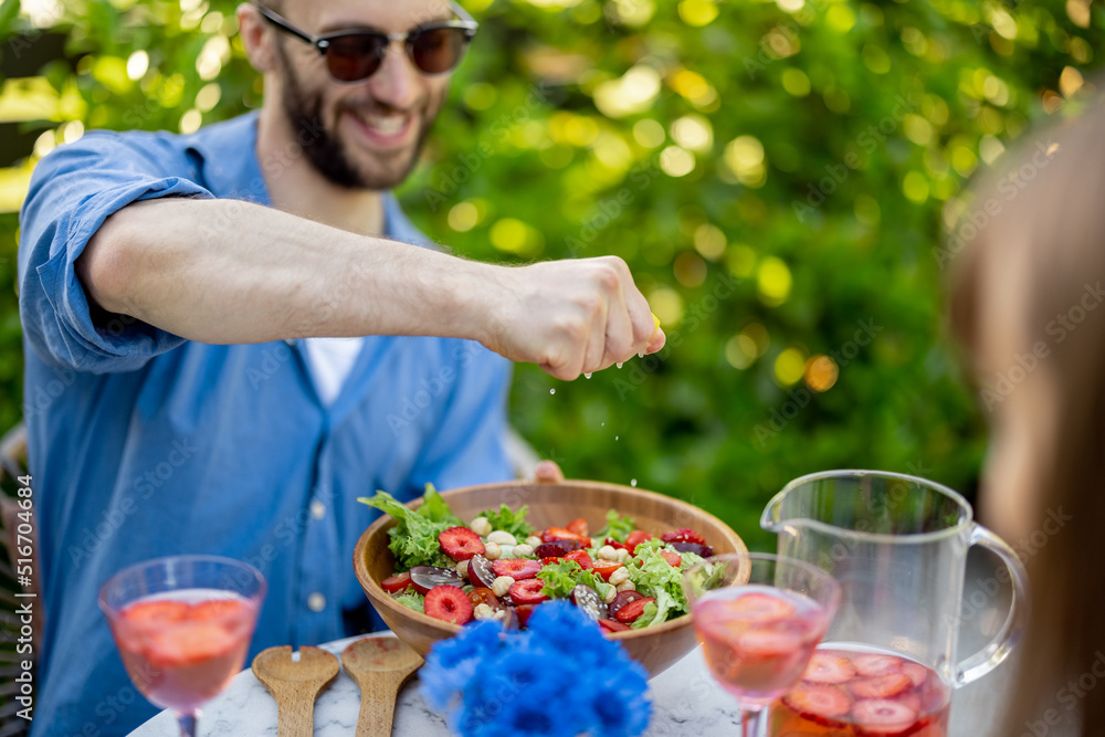 Man squeezes lemon to the salad while having lunch with woman at backyard. Young family eating healt