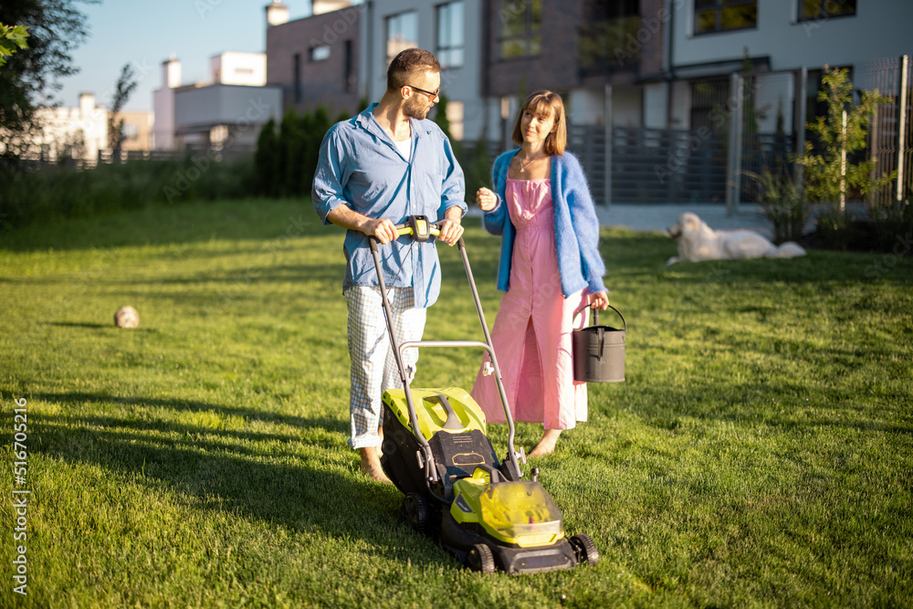 Man mows the lawn with lawn mower, spending summer time with his wife at backyard of country house. 