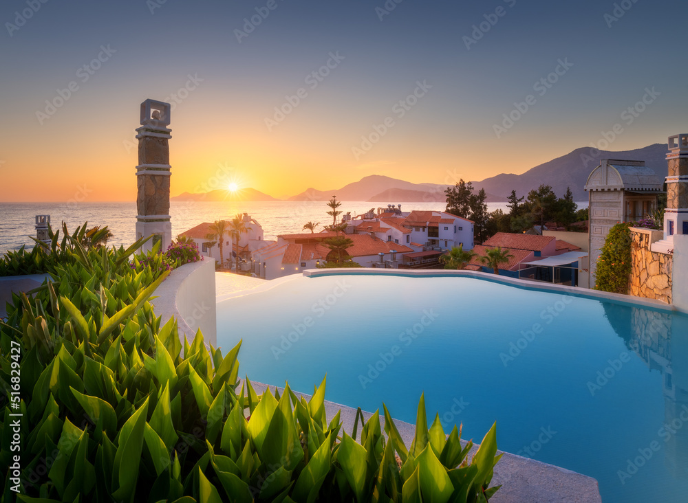 Beautiful swimming pool, green bush and orange roofs at colorful sunset. Blue water, sea coast, arch