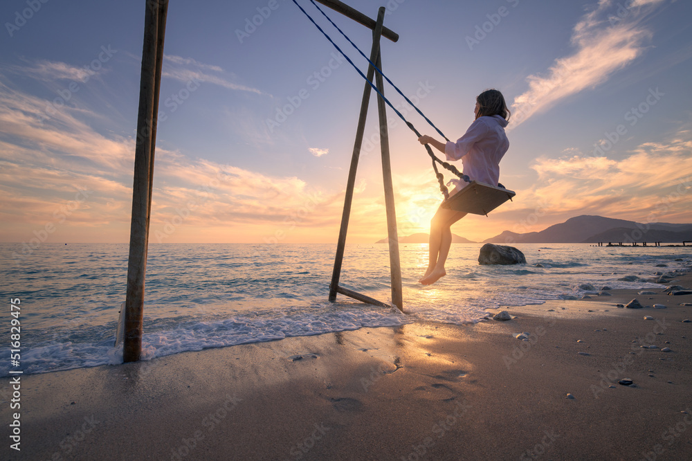 Happy young woman on wooden swing in water, beautiful blue sea with waves, sandy beach, golden sky a