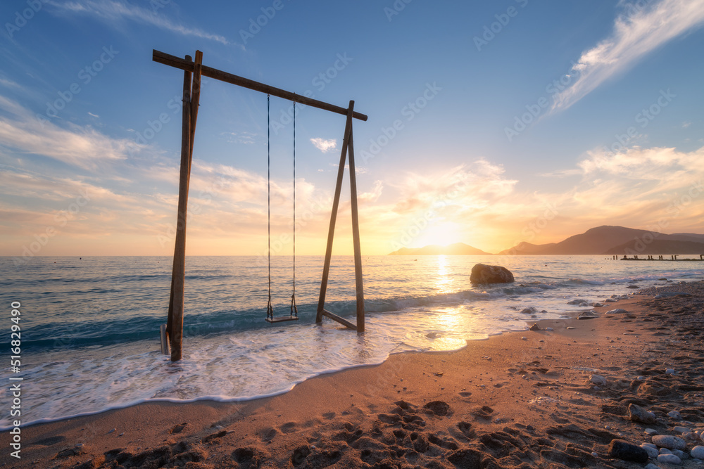 Wooden swing in beautiful blue sea with waves, sandy beach, sky with golden sunlight at sunset in su