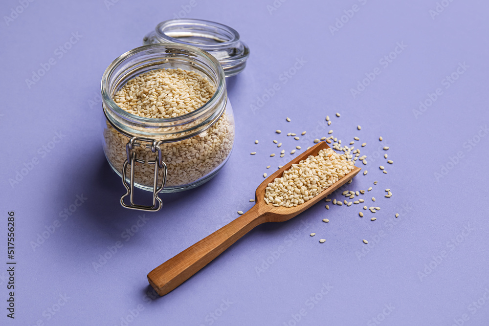 Glass jar and wooden scoop with sesame seeds on color background