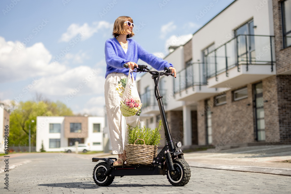 Happy stylish woman going home with fresh vegetables in mesh bag, driving on electrical scooter at r