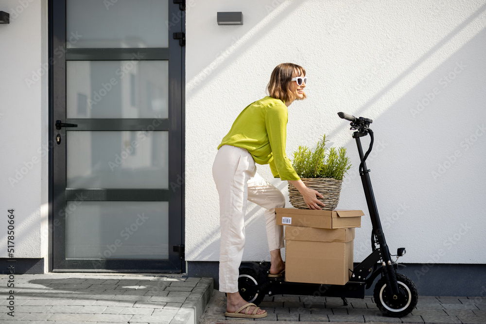 Young woman carrying basket with herbs on a porch of her house with electric scooter and parcels nea