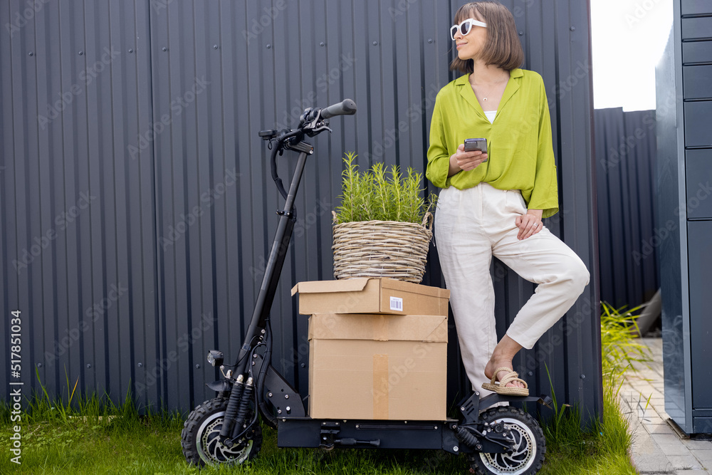 Young woman standing with phone near her electric scooter with parcels and flowerpot on gray wall ba