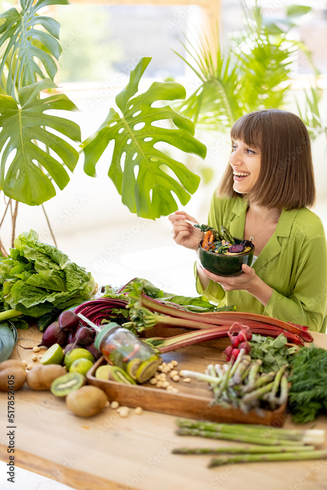 Young cheerful woman eats vegetarian bowl while sitting by the table full of fresh vegetables, fruit