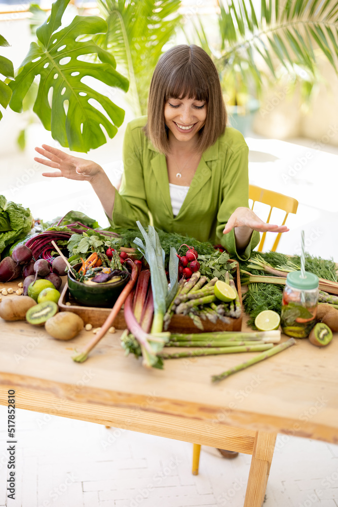 Portrait of a young cheerful sits by the table full of fresh vegetables, fruits and greens indoors w