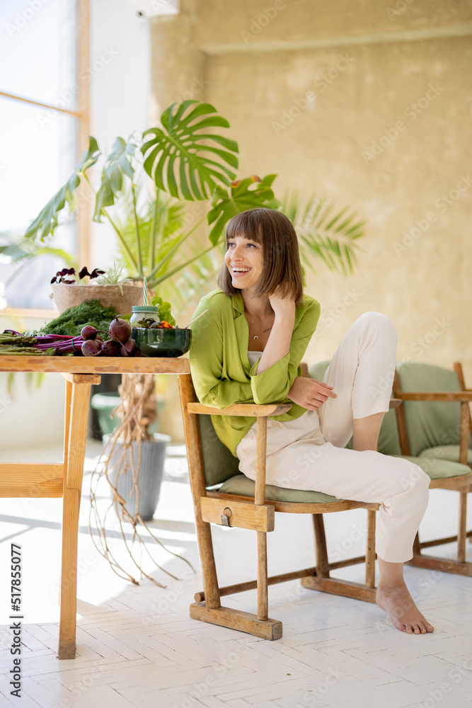 Portrait of a young cheerful woman sits relaxed on chair near the table full of fresh vegan food in 