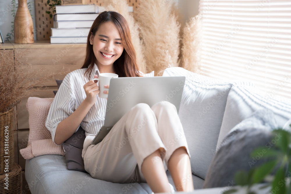 年轻的亚洲女性在家里的沙发客厅里使用笔记本电脑，在家舒适快乐地工作。