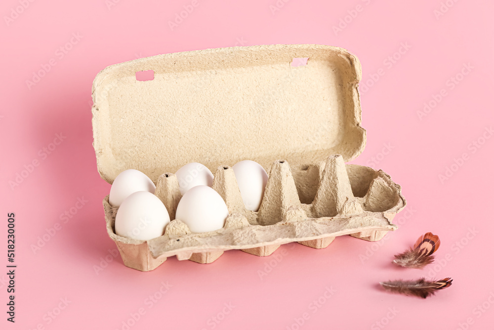 Cardboard holder with chicken eggs and feathers on pink background