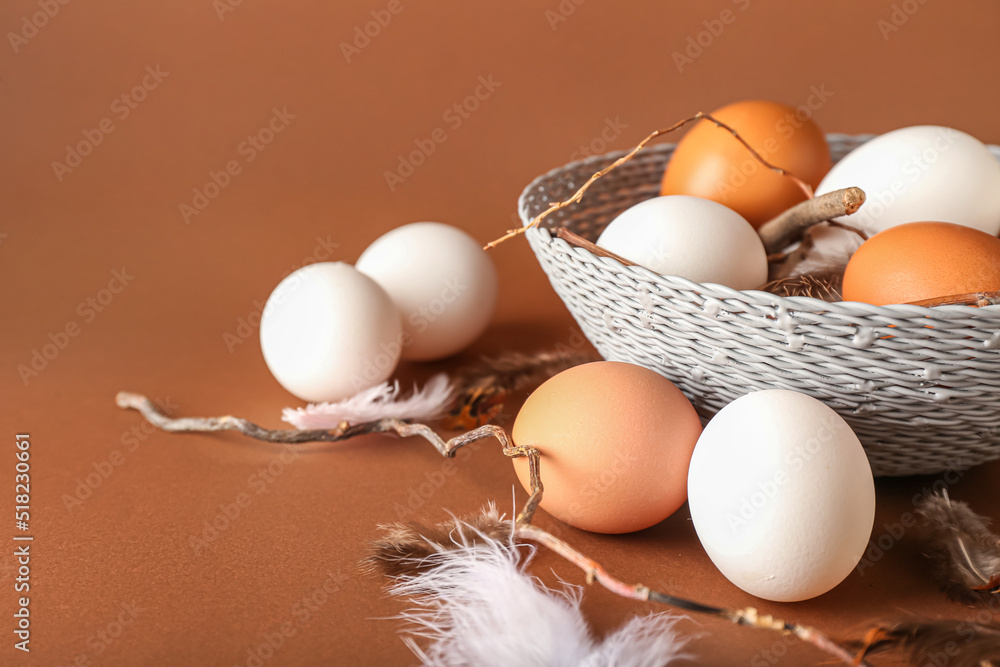 Wicker bowl with chicken eggs, feathers and tree branches on color background