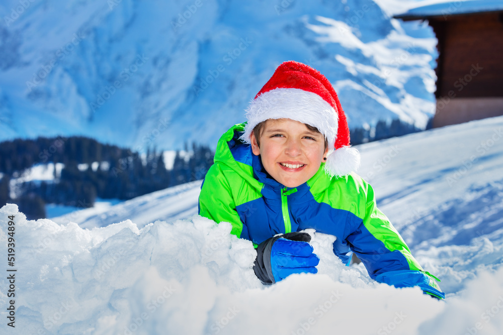 Boy with Santa hat and big smile lay in snow over mountains