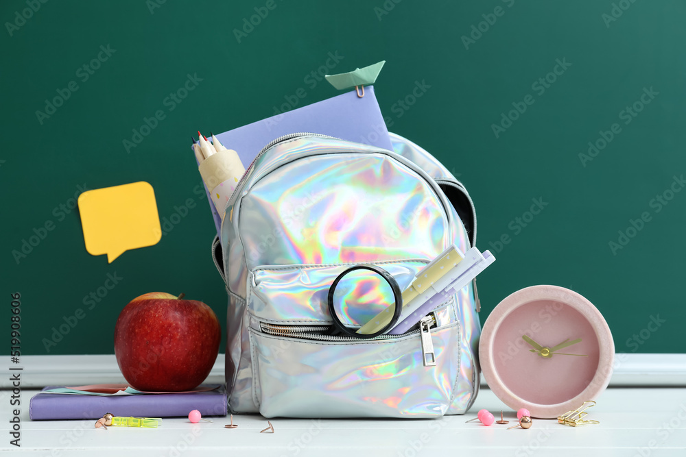 School backpack with stationery, apple and alarm clock on table near green chalkboard