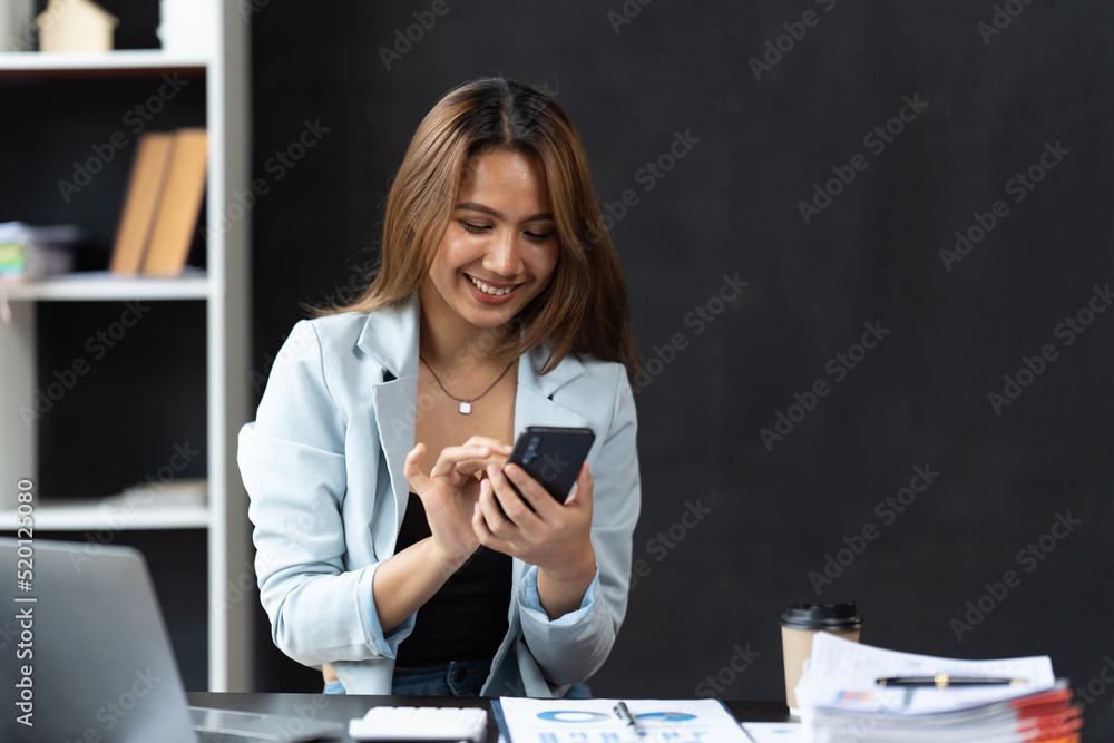 穿着正式西装的亚洲女商人在办公室里，在使用智能手机和工作时快乐而愉快。