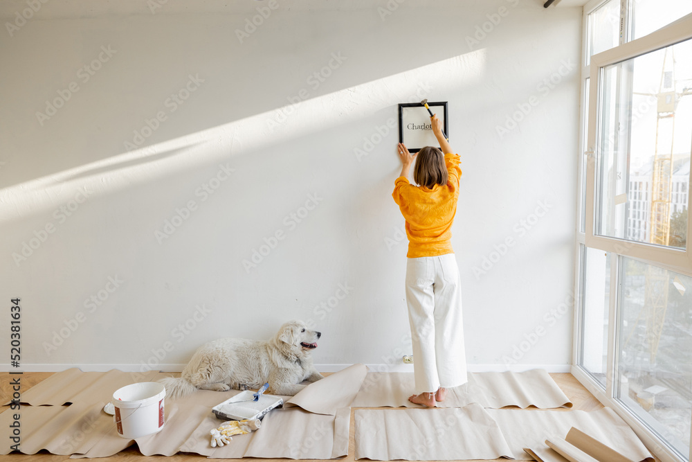 Young woman hanging picture frame in room, decorating her newly renovated apartment, stands with her