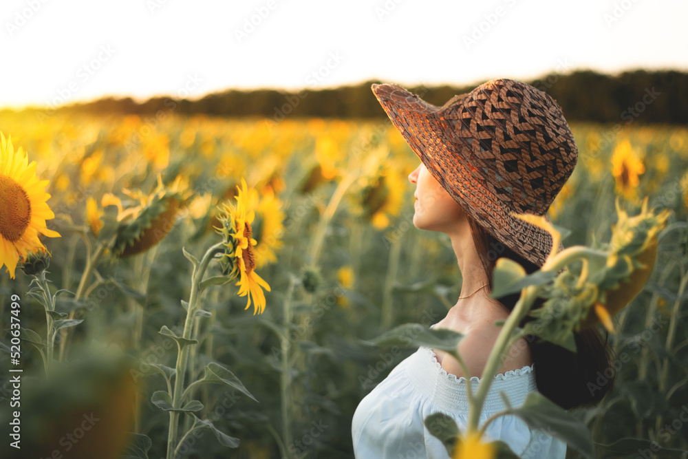 日落时，一个穿着白色连衣裙、戴着帽子的年轻女孩在向日葵地里。一个女人的肖像