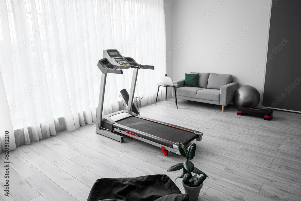 Interior of light living room with treadmill, sofa and sport equipment