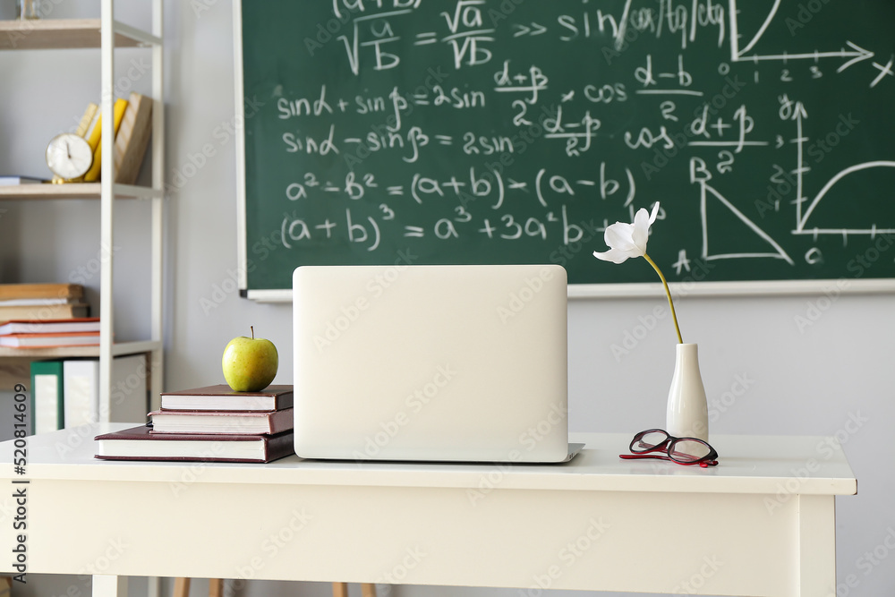 Apple, books, laptop, eyeglasses and vase with flower on table in classroom