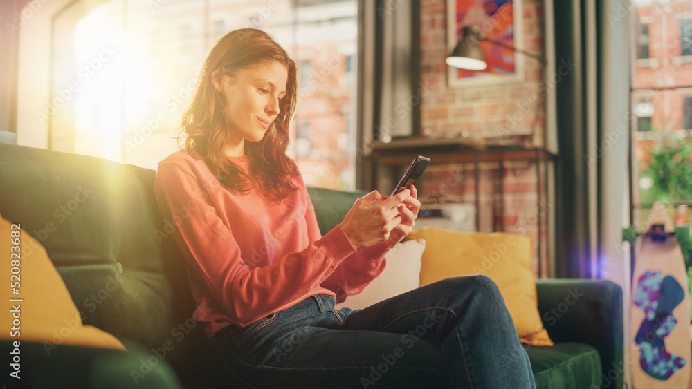 Young Beautiful Female Sitting on a Couch in Living Room, Relaxing and Using Smartphone. Creative Gi