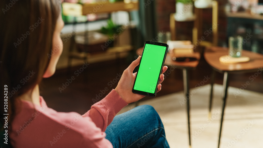 Feminine Hand Holding a Smartphone with Green Screen Mock Up Display. Female is Relaxing at Home, Wa