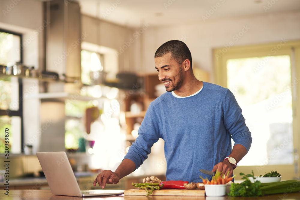 Technology has its place in the kitchen. Shot of a happy young man using a laptop while preparing a 