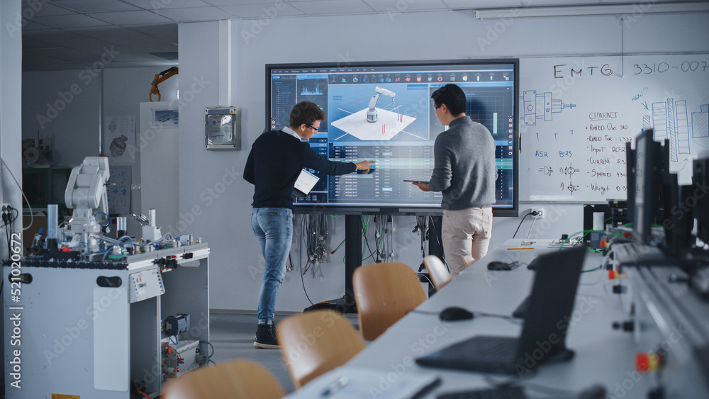 Two Diverse Male Students Standing Near Big Screen and Developing Program for Motion of Robotic Arm.