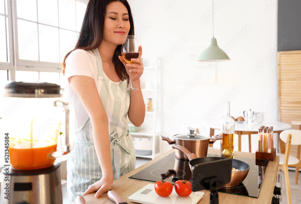 年轻的亚洲女人拿着一杯葡萄酒在厨房观看烹饪视频教程