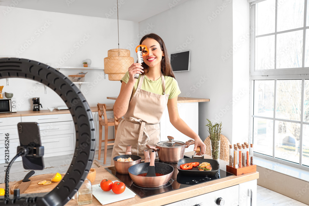 Young woman frying fish with vegetables while recording video tutorial in kitchen