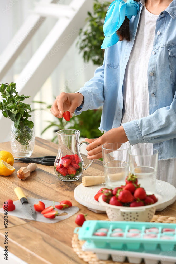 Woman putting strawberry into jug for lemonade in kitchen