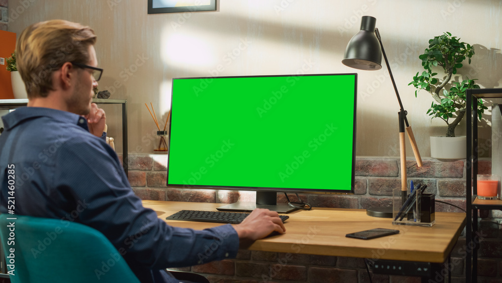 Young Handsome Man Working from Home on Desktop Computer with Green Screen Mock Up Display. Creative