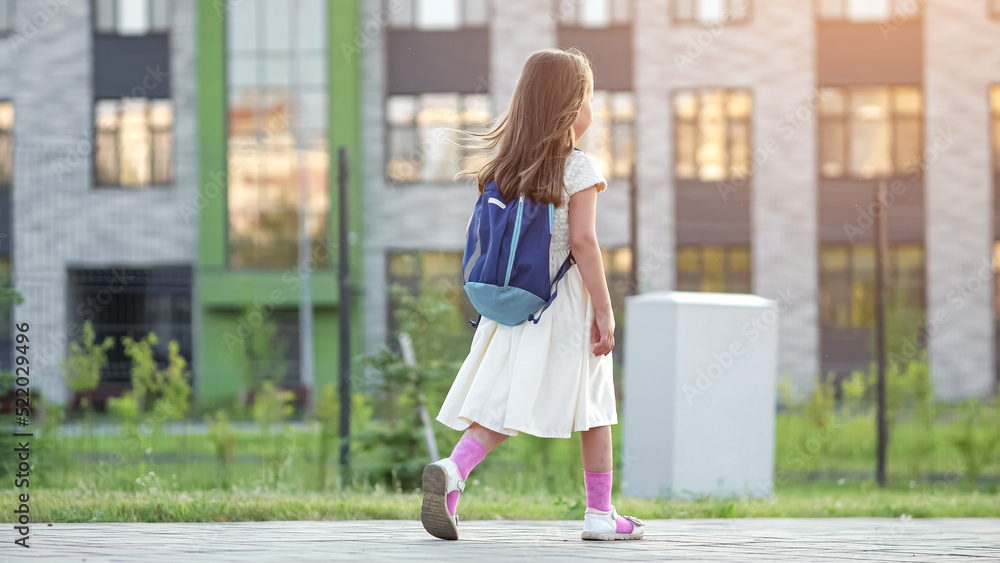 Preschooler girl with blue backpack walks past big school building. Schoolkid goes to school hopping