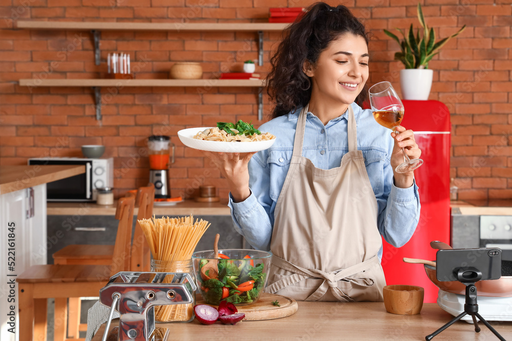 Young woman with tasty pasta and glass of wine recording cooking video class in kitchen