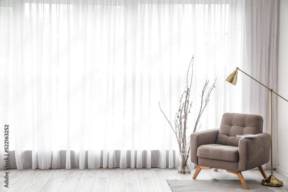 Vase with tree branches, armchair and lamp near light curtain in living room