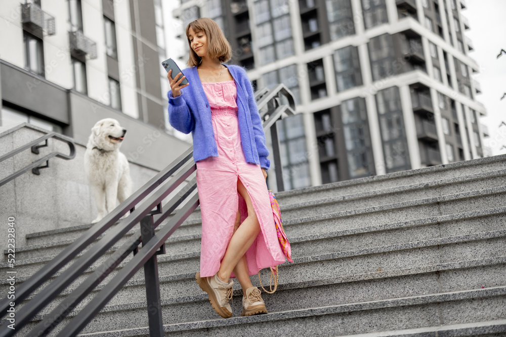 Young stylish woman dressed brightly stands with a smart phone on staircase at modern housing estate