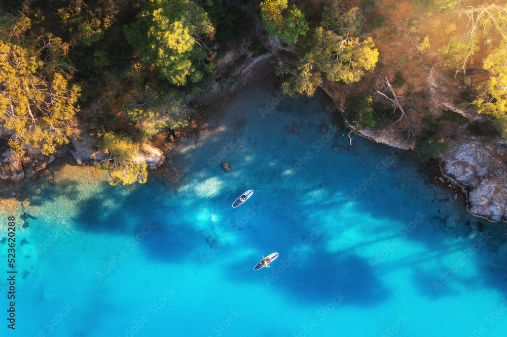 Aerial view of people on floating sup boards on blue sea, rocks, trees at sunset in summer. Blue lag