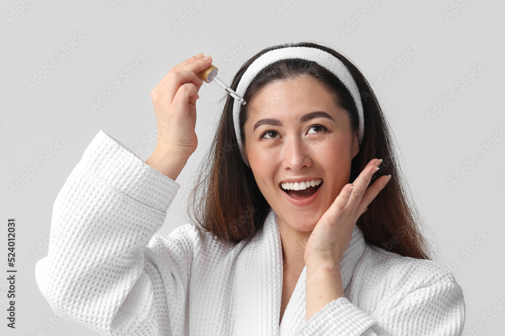 Young Asian woman with cosmetic serum on grey background