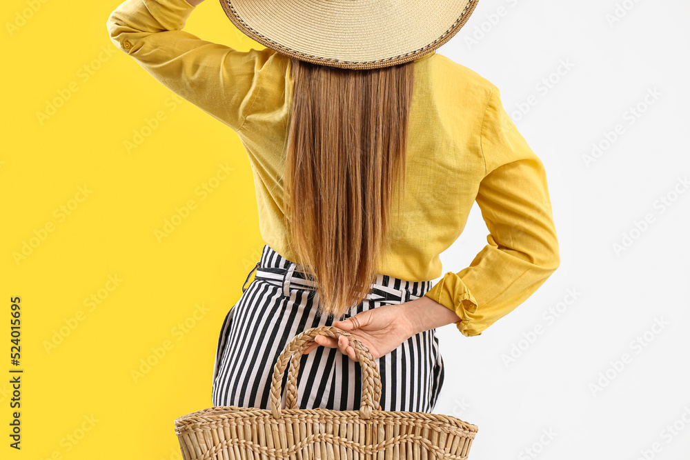 Young woman in stylish hat holding rattan handbag behind her back on color background