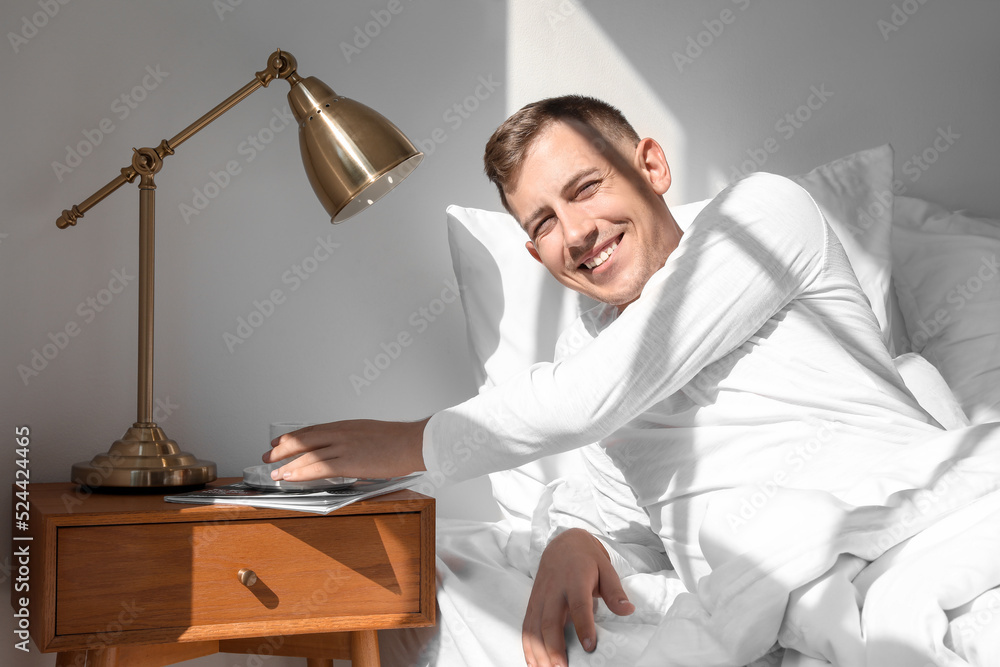 Handsome young man taking cup of coffee from table in bedroom