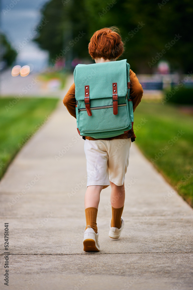 rear view of cute redhead school boy, kid with backpack walks on the street