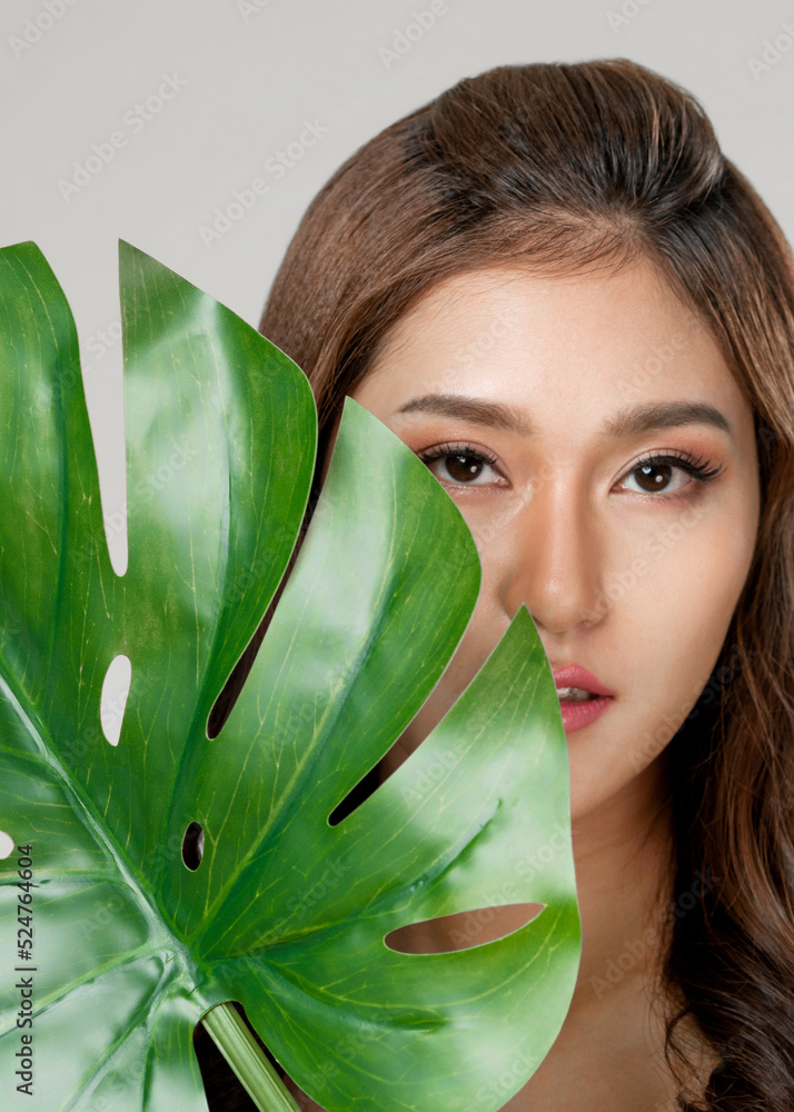 Closeup portrait of young ardent woman with healthy fair skin holding big green leaf near her face. 