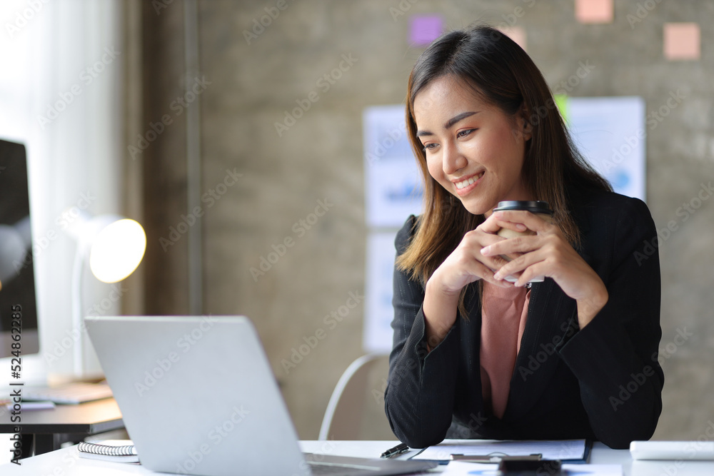 Attractive Asian businesswoman drinking coffee working on laptop in office.