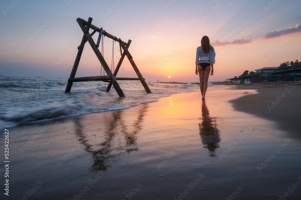 Young woman near wooden swing in water, beautiful blue sea with waves, sandy beach, reflection in wa