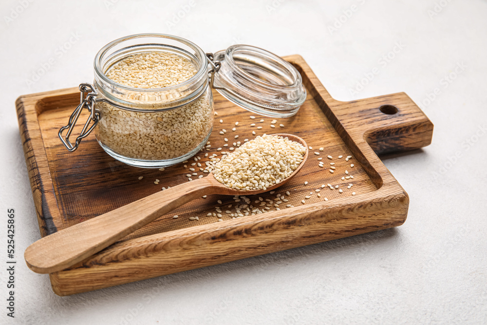 Wooden board with glass jar and spoon of sesame seeds on light background