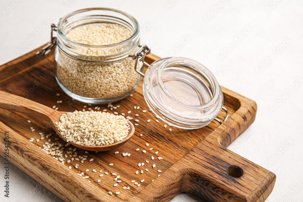 Wooden board with glass jar and spoon of sesame seeds on light background, closeup