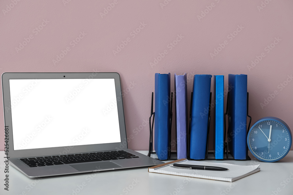 Holder with books, alarm clock and laptop on table near pink wall