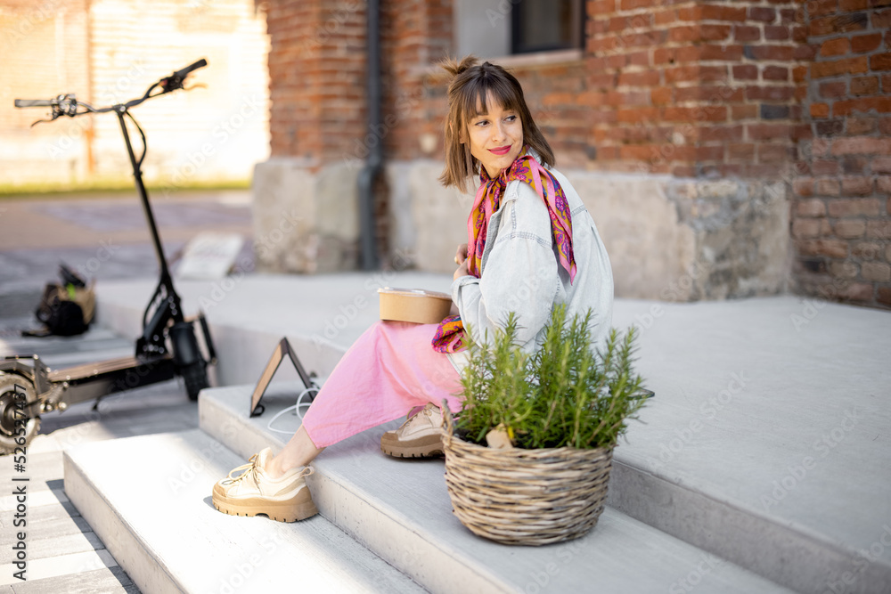 Young stylish woman has a coffee break while sitting near office building outdoors. Modern lifestyle