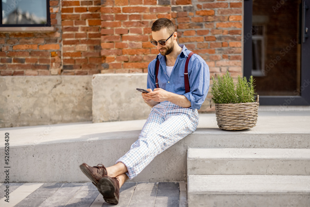 Stylish man uses smartphone while sitting on street with green plant near office building. Portrait 